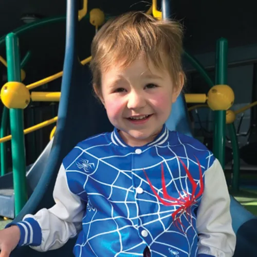 Smiling young boy in a blue spider-web jacket at a playground, supported by Small But Mighty SK